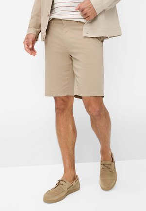 Uomo con pantaloncini beige, mocassini in suede beige abbinati, una camicia a righe e una giacca beige chiaro, davanti a uno sfondo bianco uniforme.