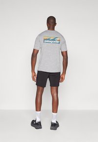 Γκρίζο κοντομάλλικο μπλουζάκι με γραφικό κύματος και λογότυπο "patagonia" στην πλάτη, συνδυασμένο με μαύρα σόρτς και αθλητικά παπούτσια.