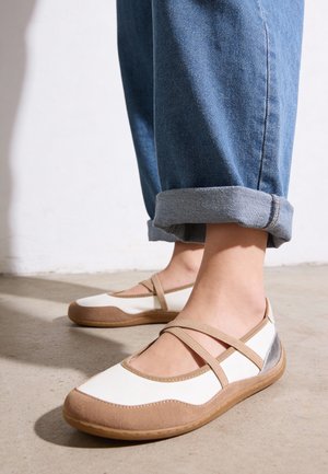 Piedi con scarpe basse beige e bianche con cinturini elastici, abbinate a jeans blu risvoltati su un pavimento in cemento.