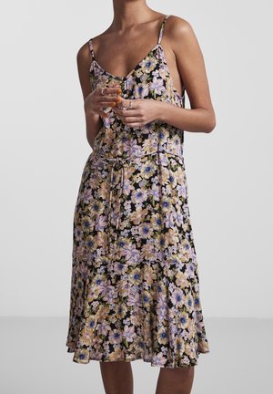 Vrouw draagt een mouwloze bloemenjurk met dunne bandjes en een gestrikte taille, staand tegen een effen lichtgrijze achtergrond.