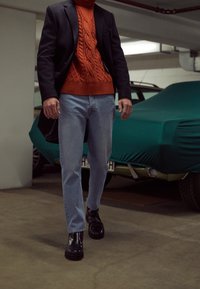 Blazer negro, suéter de cuello alto de punto trenzado naranja, jeans de mezclilla azul claro y botas de piel negras. El fondo presenta un coche cubierto.