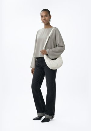 Ženska, ki nosi ohlapno sivo majico z dolgimi rokavi, črne kavbojke z zavihki, črne koničaste škornje in nosi belo torbico čez ramo z logotipom CK.