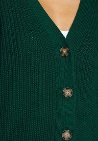 Stickad grön kofta med V-ringning, fem dekorativa knappar och texturerad ribb, som visar upp ett mjukt och varmt tyg.