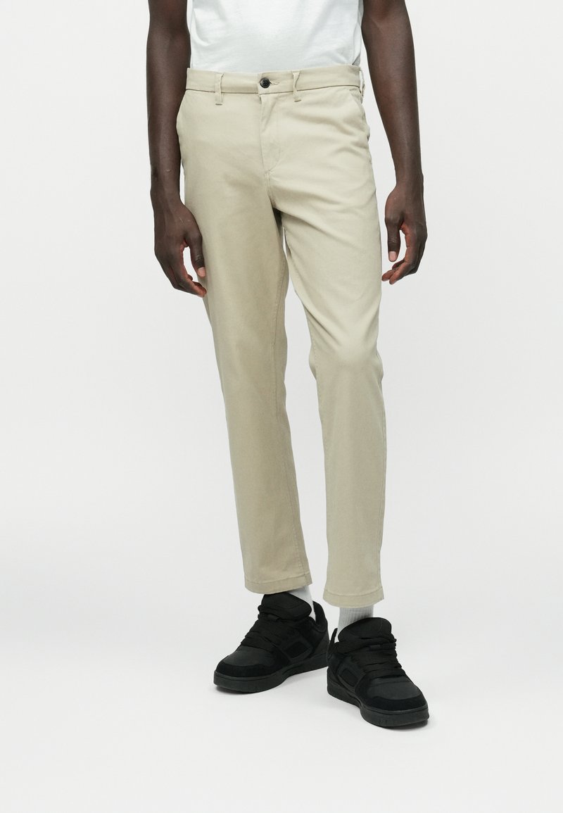 Chino en coton beige avec une coupe droite, fermeture à bouton et poches latérales. Associé à des baskets montantes noires.