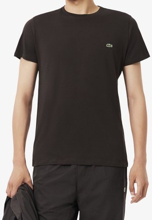 Mężczyzna noszący prostą czarną koszulkę z krótkim rękawem z małym zielonym logo krokodyla na piersi, trzymający w jednej ręce czarną kurtkę.