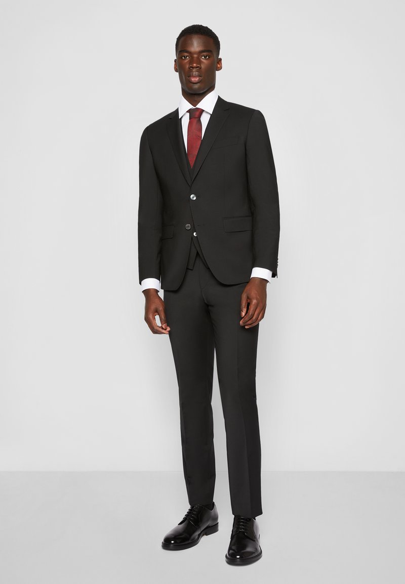 Zwarte maatpak met een op maat gemaakte pasvorm; enkel geknoopte blazer met twee knopen, gecombineerd met een wit overhemd en een dieprode stropdas. Zwarte leren schoenen.