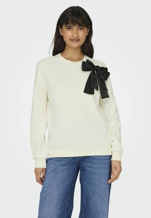 Felpa crema con un grande fiocco nero decorato con borchie sulla spalla sinistra; polsini e orlo a costine, abbinata a jeans blu.