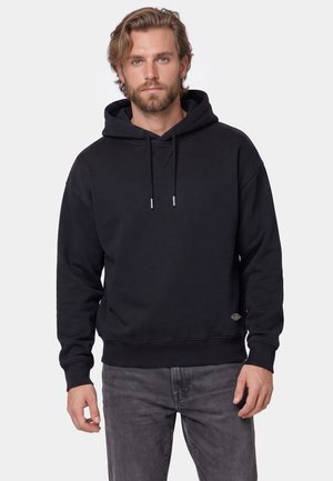 Mand med skæg iført en sort hættetrøje med snore og mørkegrå jeans stående mod en ensartet lys baggrund.