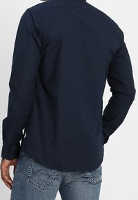 Granatowa koszula z długim rękawem wykonana z bawełny, z kołnierzykiem na guziki, dopasowanym krojem i gładką teksturą. Posiada mankiety na guziki oraz zaokrąglony dół.