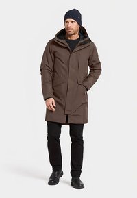 Bruine waterdichte parka met capuchon, voorzien van een ritssluiting en drukknoopsluitingen, gecombineerd met zwarte broek en een gebreide muts. Gladde textuur.