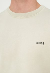 Világos bézs színű, kötött anyagból készült kereknyakú pulóver, bordázott gallérral és a mellkason egy kis fekete "BOSS" logóval.