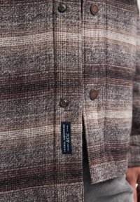 Geweven jas in bruin en grijs met een ruitenpatroon, voorzien van metalen drukknoopjes en een textiellabel aan de linkervoorzijde.