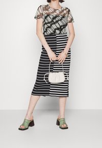 Top trasparente marrone e verde con maniche corte, abbinato a una gonna midi a righe bianco e nero. Sandali con tacco verde e borsa color crema.