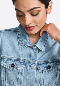 Giacca di jeans blu chiaro con colletto classico, bottoni argentati e due tasche sul petto, caratterizzata da cuciture sottili e una texture leggermente sbiadita.