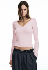 Bluza roz deschis cu mâneci lungi, decolteu în V și dantelă la marginile. Material moale, croială potrivită, combinată cu pantaloni înalți, de culoare închisă și o curea neagră.