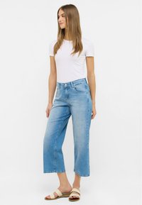 Jean bleu clair à jambe large en denim. Doté d'une taille haute, de cinq poches et d'une finition légèrement décolorée. Associé à des sandales beiges.