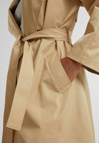 Beige trench coat featuring a tied belt, side pocket, wide sleeves, and a smooth texture. The design is simple and streamlined.