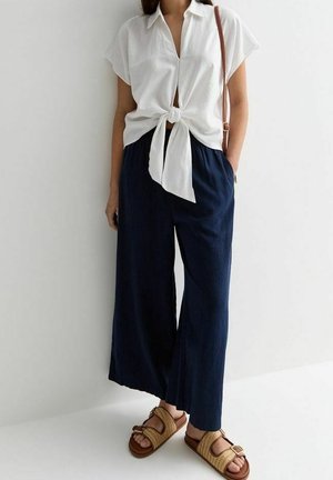Žena oblečená v bílé krátké košili s uzlem, tmavě modré široké kalhoty, hnědé sandály a hnědá kabelka přes rameno na bílém pozadí.