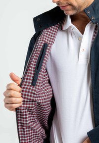 Veste navy avec fermeture éclair, dotée d'une doublure à carreaux rouges et blancs et d'une poche latérale. Le matériau semble avoir une texture douce.