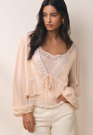 Vrouw met lang donker haar draagt een doorzichtige beige blouse met kanten rand en pofmouwen, gecombineerd met lichtgekleurde broek, staand tegen een neutrale muur.