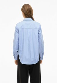 Sinivalkoraidallinen pitkähihainen paita, jossa on taakse menevä kaitale, napillinen kaulus ja hieman kaartuva helma, yhdistetty mustiin housuihin.