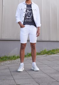 Chaqueta de mezclilla blanca sobre una camiseta gris con gráfico, combinada con pantalones cortos blancos y zapatillas grises. Superficie plana al aire libre con hierba de fondo.