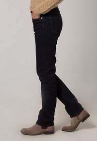 Jeans de mezclilla azul oscuro de ajuste slim, con bolsillos traseros, costuras naranjas y una textura suave. Combinados con botas de tobillo de color marrón claro.