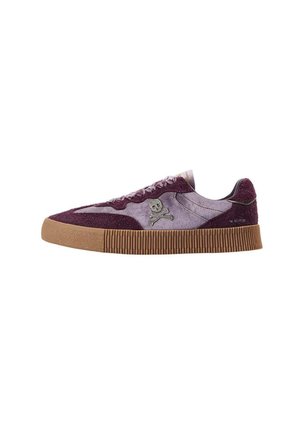 Zapatilla baja con empeine de ante púrpura, logotipo de calavera y tibias cruzadas en el lateral, suela de goma marrón acanalada y cordones planos sobre fondo blanco.