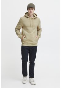 Khaki mikina s kapucňou a predným vreckom, s rebrovanými manžetami, v kombinácii s tmavými nohavicami a bielymi teniskami. Model má na sebe pletenú čiapku.