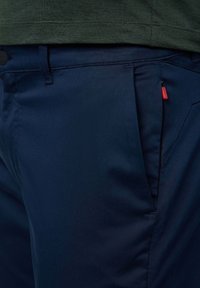 Pantalones navy con una tela suave, bolsillo lateral con tirador rojo y un diseño ajustado. Llevados con una camisa verde.