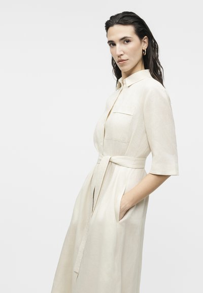 Γυναίκα με σκούρα, βρεγμένα μαλλιά φοράει κρεμ φόρεμα με ζώνη και μανίκια έως τον αγκώνα, και στέκεται με το ένα χέρι στην τσέπη σε λευκό φόντο.