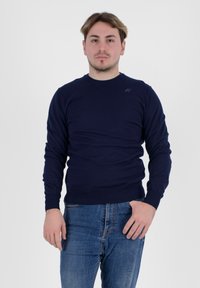 Maglione a maglia blu navy con maniche lunghe e polsini a coste, abbinato a jeans blu. Presenta un piccolo logo sul petto.