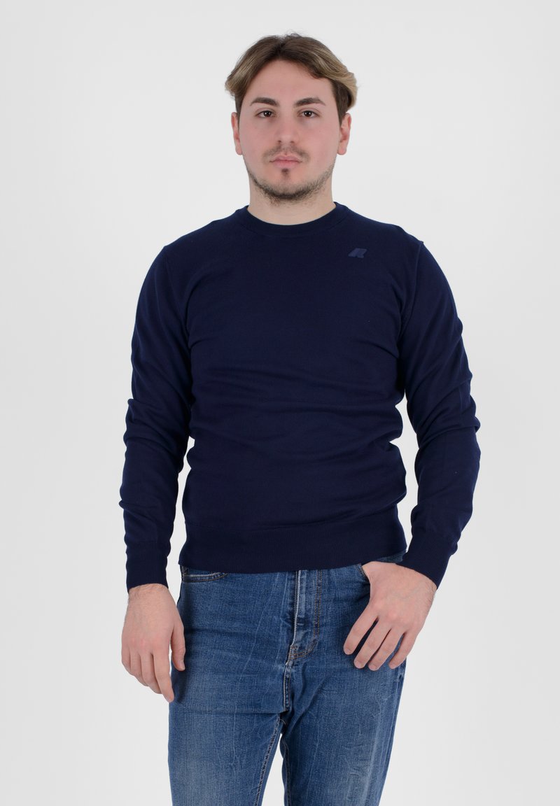 Maglione a maglia blu navy con maniche lunghe e polsini a coste, abbinato a jeans blu. Presenta un piccolo logo sul petto.