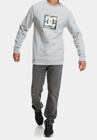 Sudadera gris con un logo de camuflaje en el frente, combinada con pantalones gris oscuro y zapatillas negras, con un corte casual y escote redondo.
