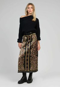 Un pull noir en maille à épaules dénudées associé à une longue jupe en satin à motifs présentant des designs dorés, violets et noirs. Bottes de combat noires.