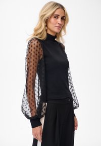 Czarna bluzka z wysokim kołnierzem i przezroczystymi, w kropki, bufkami wykonana z strukturalnej tkaniny. Posiada prążkowane mankiety i dopasowany krój.