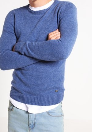 Man met over elkaar geslagen armen, draagt een gestructureerde blauwe trui met ronde hals over een wit overhemd en lichtblauwe jeans, tegen een effen achtergrond.