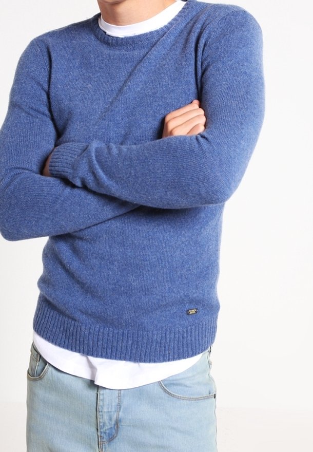 Homme les bras croisés portant un pull à col rond bleu texturé sur une chemise blanche et un jean bleu clair, sur fond uni.