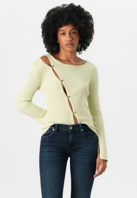 Lys gul ribbet genser med et unikt utsnitt på siden, prydet med gullknapper og vid hals, kombinert med mørkeblå jeans.