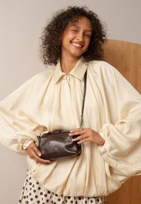 Donna sorridente con capelli ricci che indossa una giacca beige oversize con zip e una gonna a pois, mentre tiene una piccola borsa a tracolla in pelle marrone scuro.