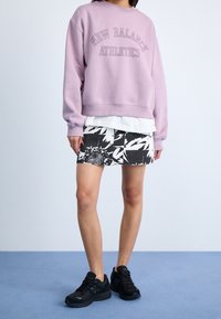 Sudadera lila con texto "NEW BALANCE ATHLETICS", camisa blanca superpuesta, falda con estampado en blanco y negro, y zapatillas deportivas negras.
