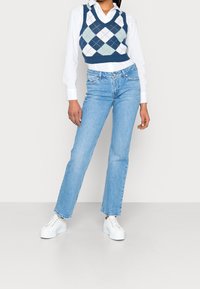 Veste en maille à losanges bleue et blanche portée sur une chemise blanche à col, associée à un jean denim bleu clair à jambes droites et des baskets blanches.