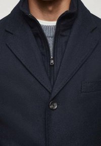 Man in een donkerblauwe jas over een grijze vest met ritssluiting en een witte crewneckshirt, wat gelaagde winterkleding toont.