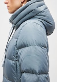 Blauwe gewatteerde jas met een hoge, opgevouwen capuchon, doorgestikt design, zachte textuur, met de merknaam "Marc O'Polo" op de linkermouw.