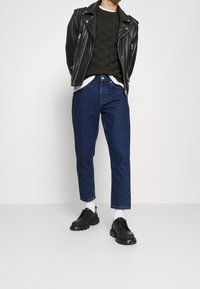 Zwarte leren motorjack over een donkergroene gestructureerde trui, gecombineerd met donkerblauwe recht gesneden jeans en zwarte stevige veterschoenen.
