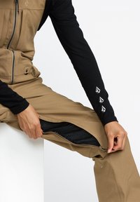 Salopette beige avec une ouverture zippée, associée à un t-shirt noir à manches longues avec trois accents de logo blancs sur la manche.