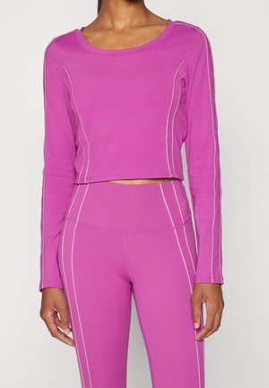 Nainen, jolla kirkkaan pinkki pitkähihainen crop-top ja korkeavyötäröiset leggingsit, joissa vaaleanpunaiset sauman yksityiskohdat, yksivärisellä taustalla.