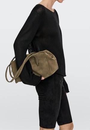 Person iført sort langærmet top og shorts, der holder en stor olivengrøn ruskindshåndtaske under armen mod en ensfarvet baggrund.