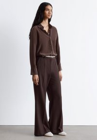 Woman standing wearing a dark brown button-up shirt and wide-leg trousers, beige belt, silver necklace, and white shoes against a plain wall.