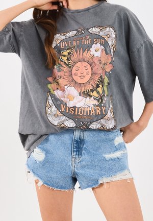 Osoba nosząca szarą, oversize koszulkę z grafiką słońca, kwiatów i motyli oraz tekstem "Live by the Sun Visionary" w połączeniu z podartymi szortami dżinsowymi.
